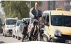 Daniel Craig in een grijs pak op een motorfiets in druk stadsverkeer, naast een gele taxi.