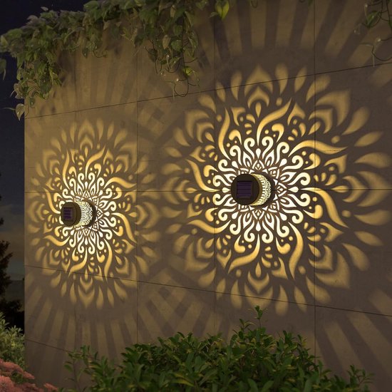 Twee zwarte wandlampen met een open, gouden mandala-patroon. Ze projecteren een stralend licht op de muur en omliggende begroeiing.