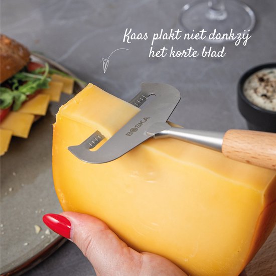 Een gele Boska kaasschaaf met een houten handvat schaaft een stuk kaas, tekst: Kaas plakt niet dankzij het korte blad