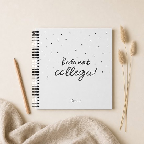 Wit notitieboek met de tekst 'Bedankt collega!' in kalligrafie en kleine zwarte stippen. Een potlood ligt links, gedroogde grassprieten rechts.