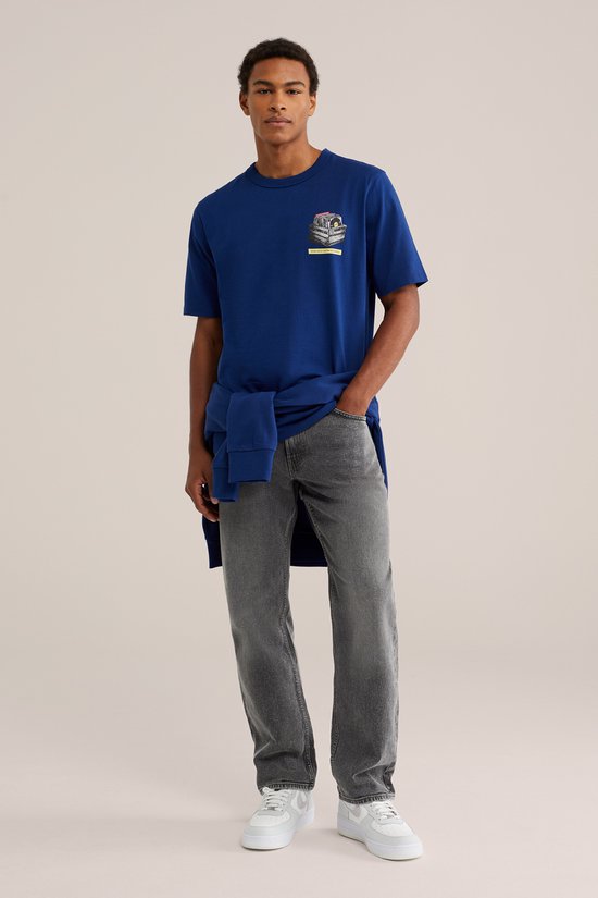 Model staat frontaal, draagt blauwe T-shirt met print en grijze jeans. Blauwe trui losjes om de nek gebonden.