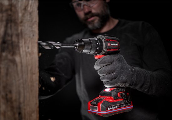 Een man met handschoenen boort in een houten plank met een rode en zwarte Einhell accuboormachine.