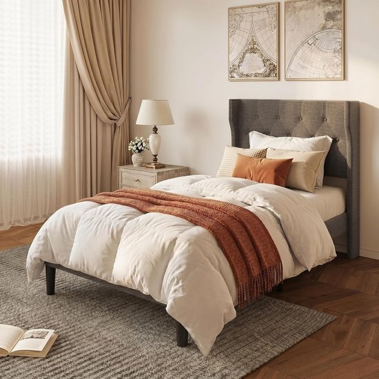 Lit simple avec couette blanche, jeté orange et tête de lit rembourrée grise, à côté d'une table de chevet avec lampe.