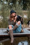 Model in bruine trenchcoat, blauwe jeans, bruine laarzen en zwarte hoed, met verrekijker.