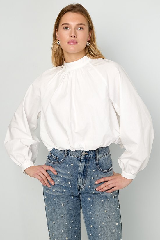 Model in een spijkerbroek met pareltjes en een witte blouse met hoge hals, ballonmouwen en strik op de rug.