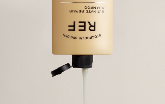 Crèmeachtige shampoo druipt uit een beige fles met zwarte dop. Tekst: REF STOCKHOLM SWEDEN ULTIMATE REPAIR SHAMPOO.