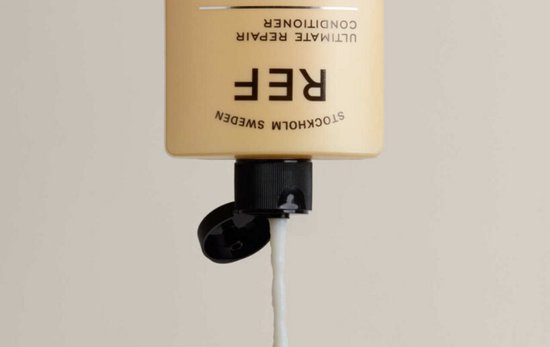 Crèmeachtige conditioner druipt uit een beige fles met zwarte dop. Tekst: REF STOCKHOLM SWEDEN ULTIMATE REPAIR CONDITIONER.