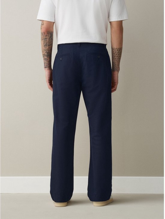 Achterzijde van donkerblauwe chino met twee achterzakken, gedragen door een man met tatoeages.