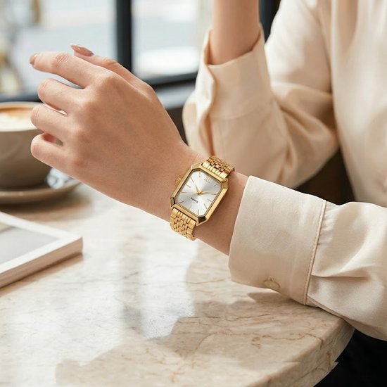 Gouden rechthoekig analoog horloge met witte wijzerplaat en Romeinse cijfers, gedragen om een pols, naast een kop koffie.
