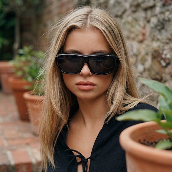 Een blonde vrouw met lang haar draagt een zwarte overzetzonnebril.