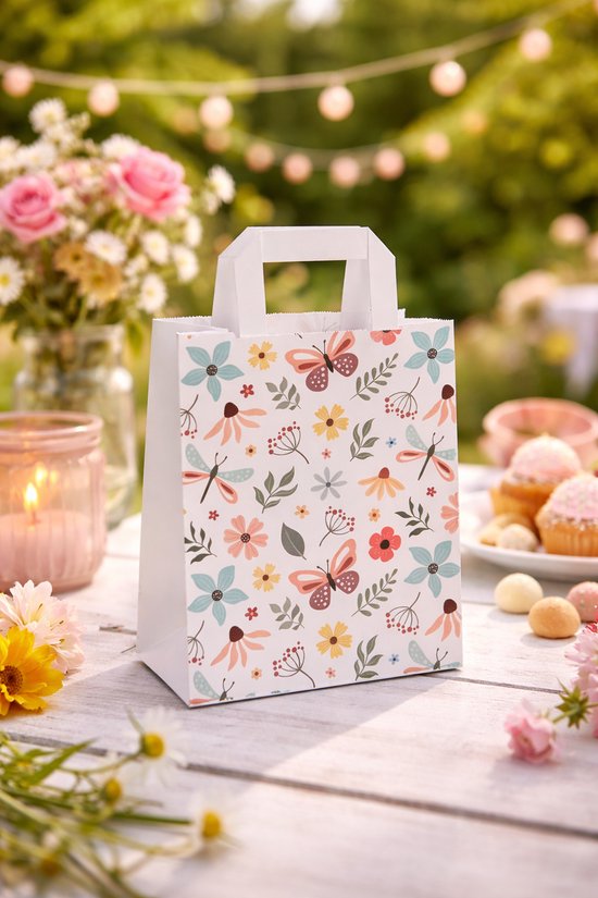 Papieren tas met vlinders, libellen en bloemenprint op een tafel in de tuin, met kaars en cupcakes.