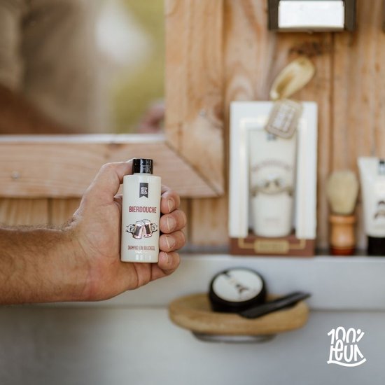 Hand houdt flesje 'BIERdouche SHAMPOO EN DOUCHEGEL' van 100% LEUK, met andere producten op achtergrond.