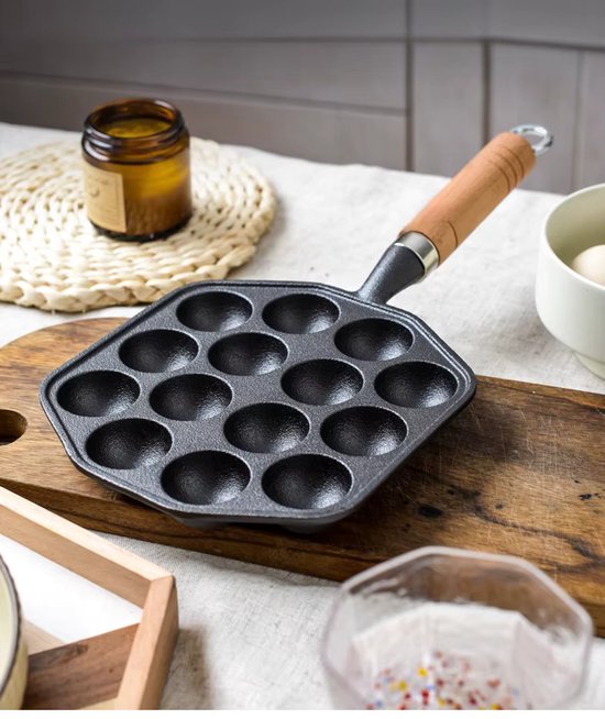 Gietijzeren poffertjespan met 14 holtes en een houten handvat, staand op een houten plank.