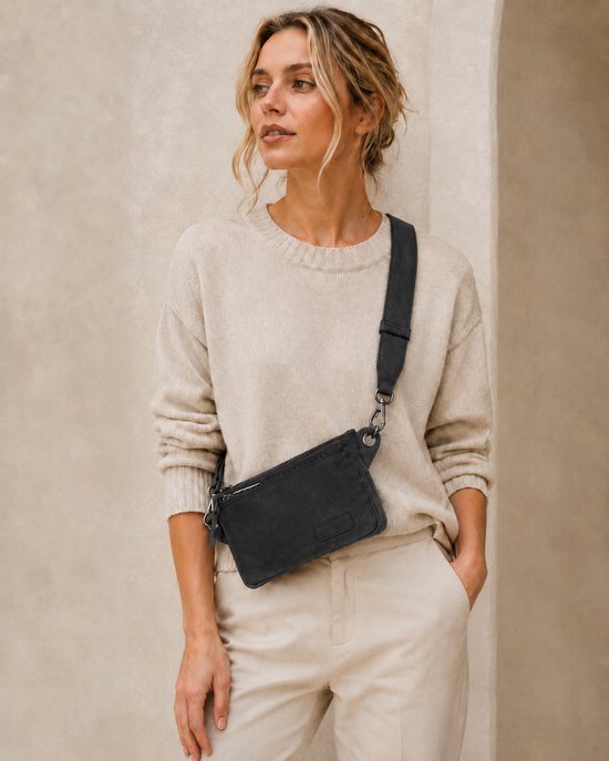 Femme aux cheveux blonds, portant un pull beige et un pantalon beige. Elle porte un petit sac banane noir en bandoulière sur son corps.