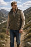 Man met olijfgroene gewatteerde bodywarmer, lichte trui en spijkerbroek, wandelend op een bergpad.