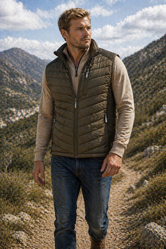 Man met olijfgroene gewatteerde bodywarmer, lichte trui en spijkerbroek, wandelend op een bergpad.
