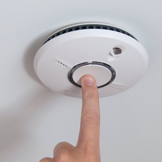 Vinger drukt op de ronde zilveren testknop van de witte rookmelder aan het plafond.