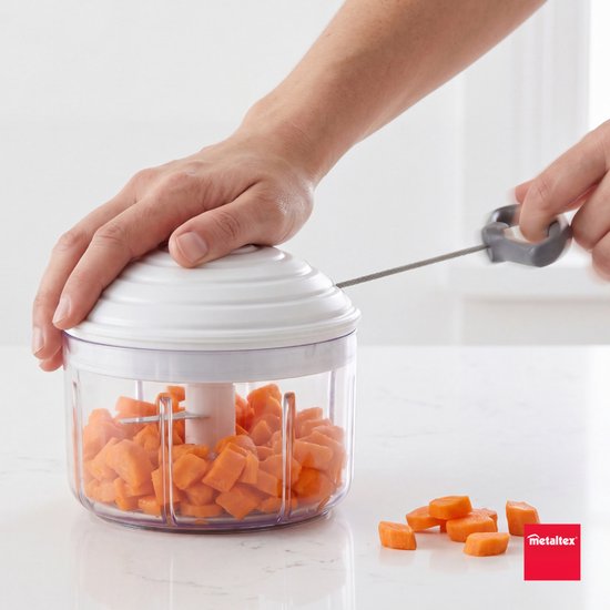 Een hand trekt aan de grijze hendel van de witte keukenmachine, gevuld met blokjes wortel.
