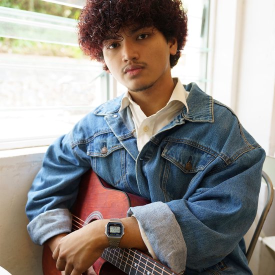 Jonge man met gekruld haar en sproeten, draagt een spijkerjack en een zilverkleurig Casio horloge om zijn linker pols, terwijl hij een akoestische gitaar vasthoudt.