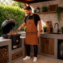 Man met bruin kunstleren schort met twee zakken en bruine leren accenten, terwijl hij kookt op een zwarte barbecue.
