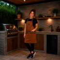 Vrouw met bruin kunstleren schort met twee zakken en bruine leren accenten, staand voor een outdoor keuken met barbecue.