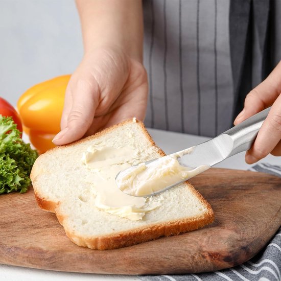 Een hand smeert boter op een stuk toast met een zilverkleurig botermesje.
