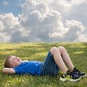 Jongen ligt op gras met Pikachu-sneakers, met blauwe lucht en wolken op de achtergrond.