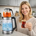 Een lachende vrouw met blond haar houdt een glas rode thee vast. Naast haar staat een glazen waterkoker met een theepot bovenop, gevuld met amberkleurige thee. De waterkoker heeft blauwe LED-verlichting.