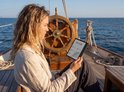Een vrouw met krullend blond haar leest een PocketBook Era Lite op de boeg van een schip.