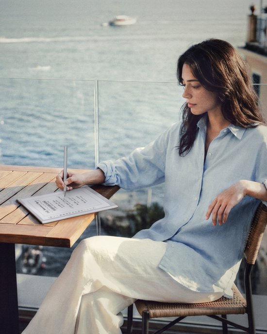 Een vrouw met een lichtblauwe blouse gebruikt een styluspen op een BOOX e-reader aan een tafel met uitzicht op zee.