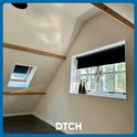 Chambre mansardée avec deux fenêtres de toit inclinées et une fenêtre normale, toutes deux avec des stores enrouleurs noirs.
