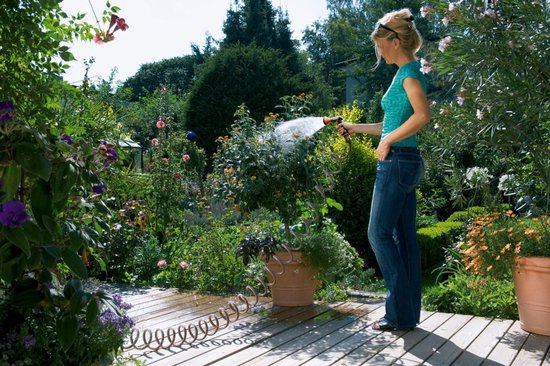 Vrouw met blauwe spijkerbroek en turkooise top besproeit planten met tuinslang in een weelderige tuin.