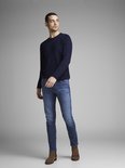Man in donkerblauwe trui en blauwe jeans, staand opzij.
