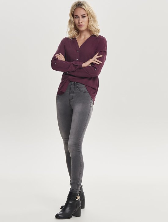 Vrouw staand in grijze skinny jeans en bordeauxrode blouse.