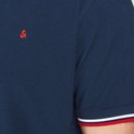 Close-up van het rode logo op de borst van een donkerblauwe polo met rood-wit gestreepte mouwboord.
