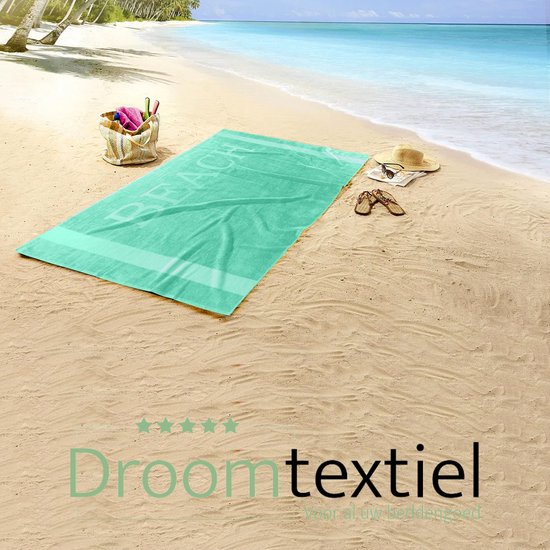 Mintgroen Droomtextiel strandlaken met "BEACH" erop, liggend op het zand bij de oceaan. Naast een mand met handdoeken en een hoed.