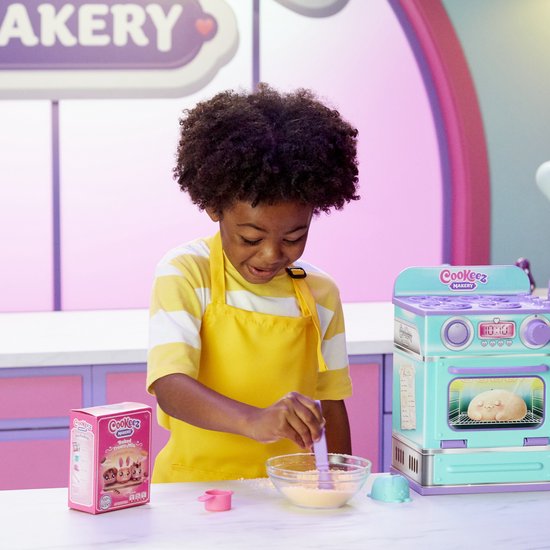 Jongen met een gele schort die een paarse lepel in een kom met poeder roert. Naast hem staat een turkooise en paarse speelgoedoven en een doos van "Cookeez Makery".