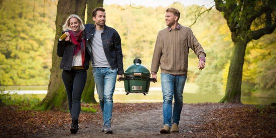 Drie personen die een donkergroene Big Green Egg barbecue dragen in een bosrijke omgeving.