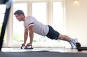 Een man in een wit shirt en zwarte korte broek doet een push-up met zwarte opdruksteunen op een mat, bij een raam.