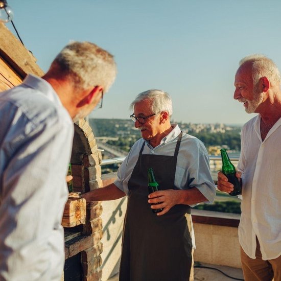 Drie mannen met bierflesjes bij een barbecue, één draagt een schort.