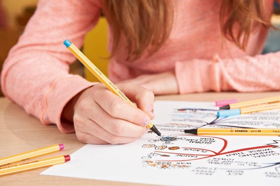 Dichtbij een hand van een meisje met een oranje fineliner die tekent op een wit papier met tekeningen. Meer fineliners liggen op tafel.