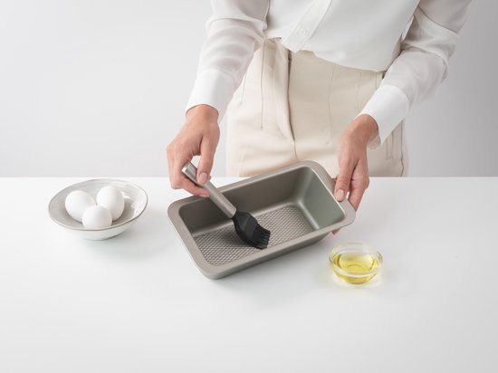 Mains graissant un moule à pain avec un petit pinceau de pâtisserie argenté à poils noirs.