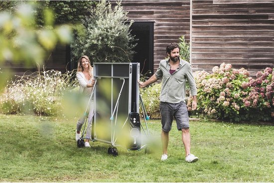 Man en vrouw duwen een blauw-grijze tafeltennistafel met wielen door een tuin.