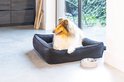 Collie in een donkergrijze hondenmand met ribbelstof, naast een voerbak.