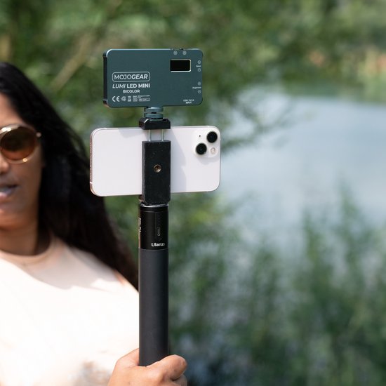 Een vrouw met zonnebril houdt een smartphone met MOJOGEAR LUMI LED MINI BICOLOR lamp vast. De telefoon is aan een selfiestick bevestigd.