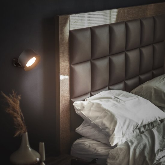 Zwarte wandlamp met warme verlichting, bevestigd aan een muur naast een bed.