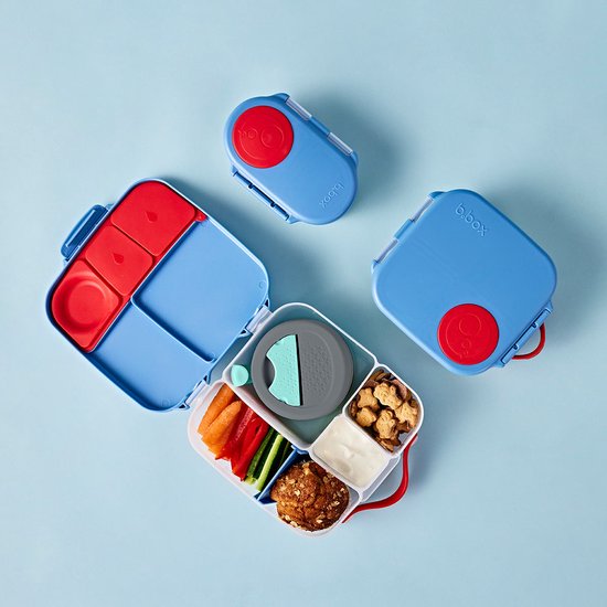 Blauwe lunchboxen, opengeklapt met rode en blauwe vakken, met eten. Een zilveren drinkfles met rode dop.