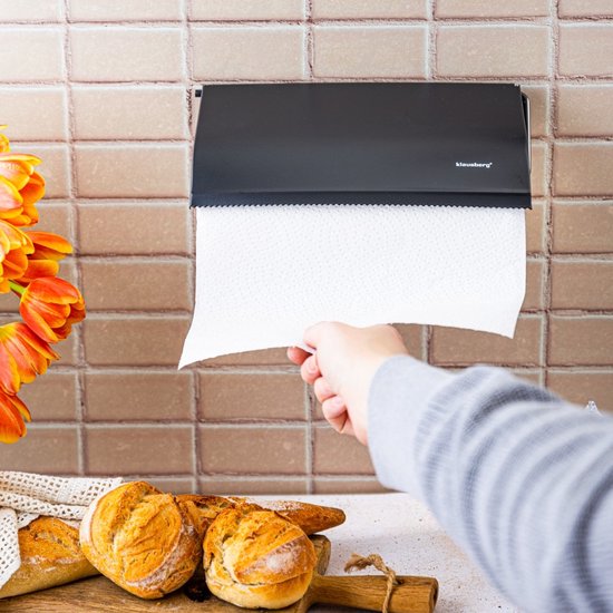 Een hand trekt keukenpapier uit een zwarte keukenrolhouder aan een betegelde muur.