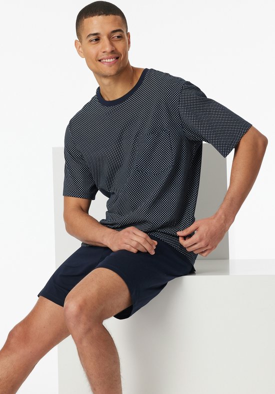Man zit op een witte kubus, draagt een donkerblauwe pyjamaset met een ruitjespatroon op het shirt.