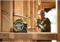Vrouw met bril meet met gele Stanley FatMax rolbandmaat in een houten structuur.